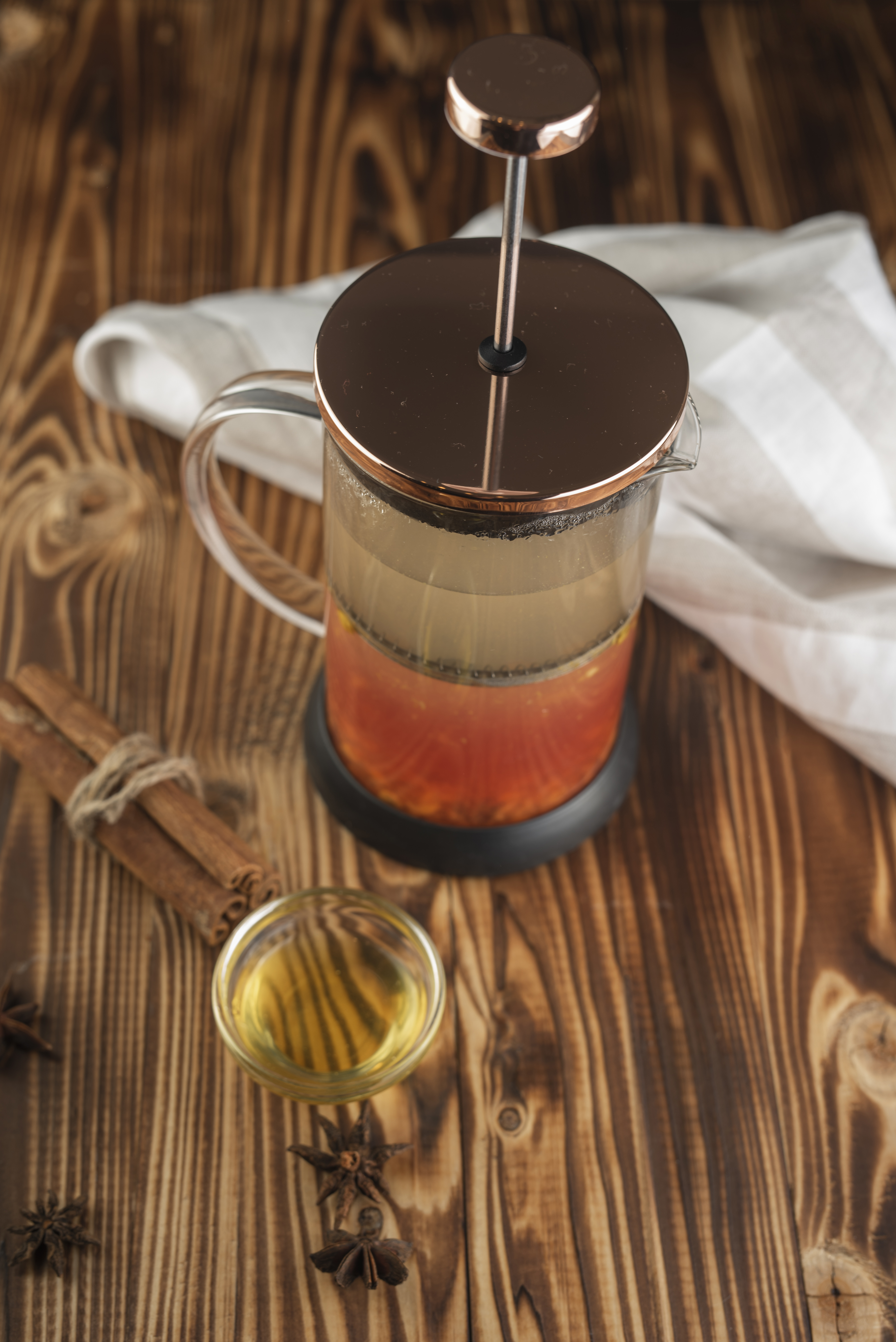 Infusion de cascara aux reflets ambrés dans une cafetière à piston en verre au couvercle cuivré, posée sur une table en bois rustique à côté d'un petit bol de liquide doré, de bâtons de cannelle ficelés et d'étoiles de badiane.